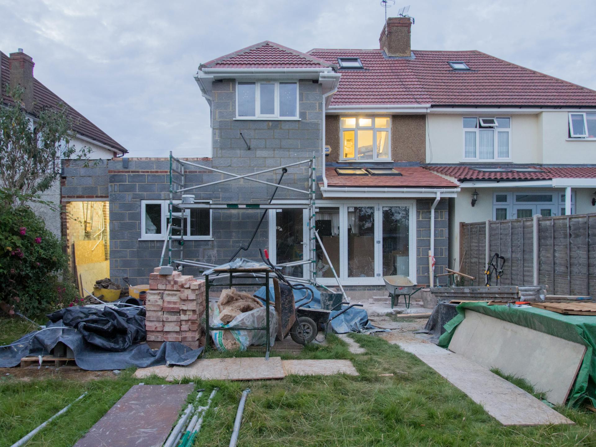 Rear view of a semi-detached house during extension works, grey blockwork and scaffolding in place, with bricks, tools and wheelbarrows across the garden.