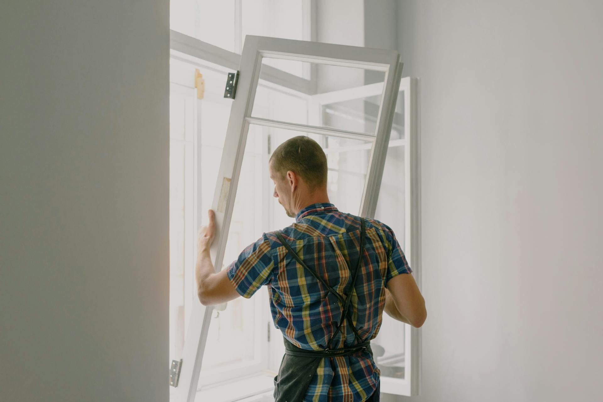 Tradesperson in a plaid shirt installing a window frame inside a bright room with open casements.e 