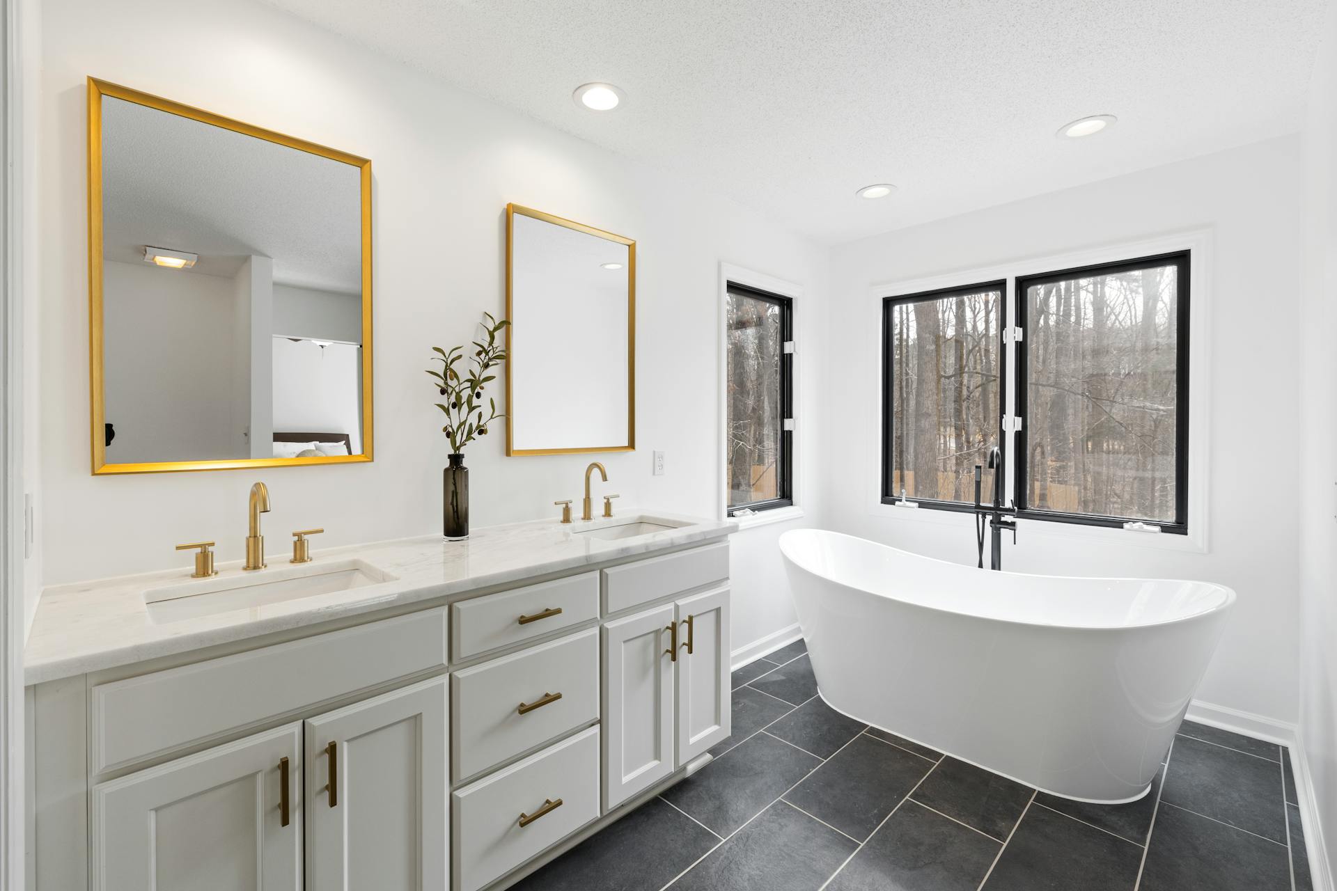Bright modern bathroom with freestanding white bath, double vanity, gold taps and mirrors, and large black-framed windows.